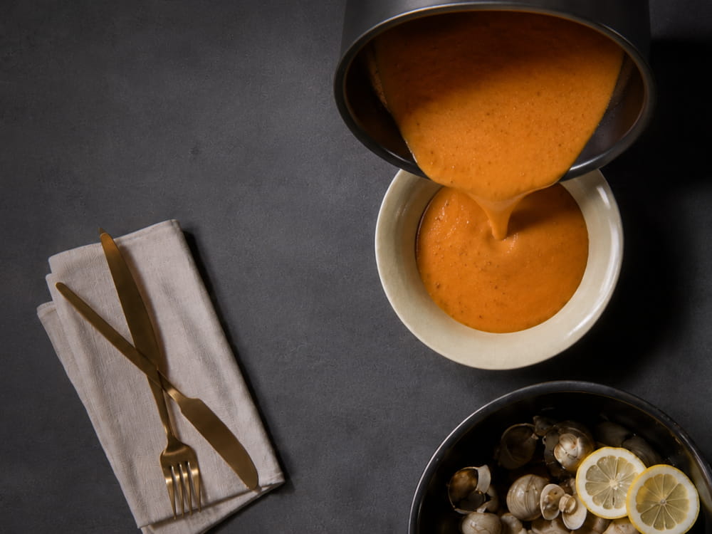 Chef pouring orange liquid from saucepan into a bowl, next to a bowl of clams
